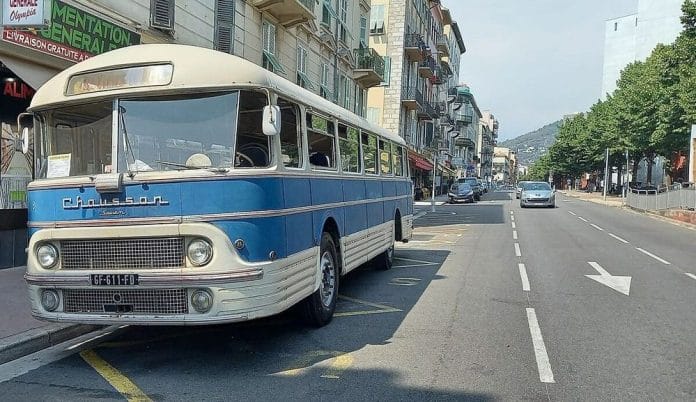 L'autocar sur une place de parking à Nice à la vue des passants. Il est dans un style rétro blanc avec une grande bande bleu claire en son centre le long de la carrosserie.