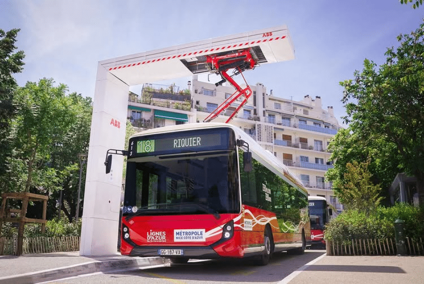 Le bus électrique en circulation à Nice de couleur blanche sur le toit et rouge pour la carrosserie en train de se recharger à une station avec un bras mécanique sur le dessus.