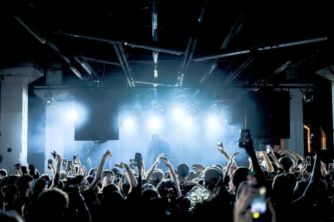 Un DJ devant une salle qui lève les bras avec des jeux de lumière blanche.