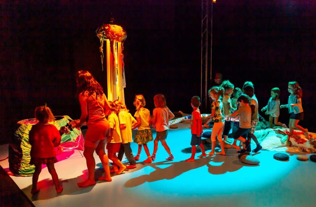 Des enfants jouent sur scène avec une animatrice dans une salle à l'ambiance très colorée par des jeux de lumière.