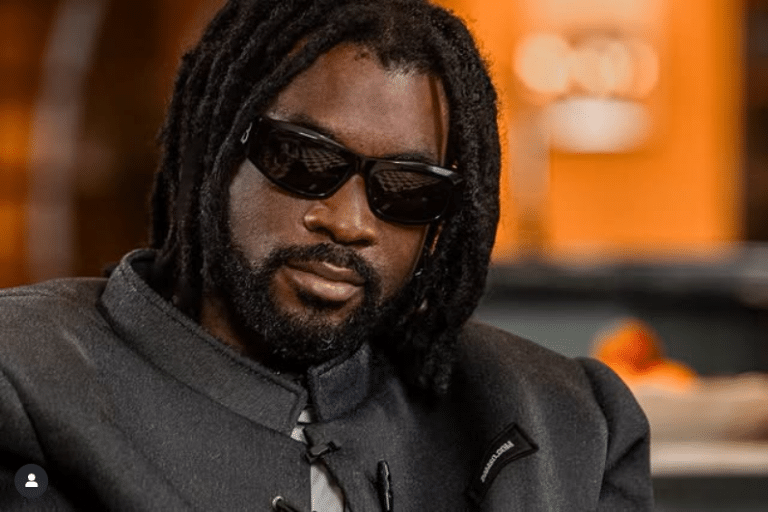Le rappeur Belge Damso avec des lunettes de soleil prend la pause habillé en noir.