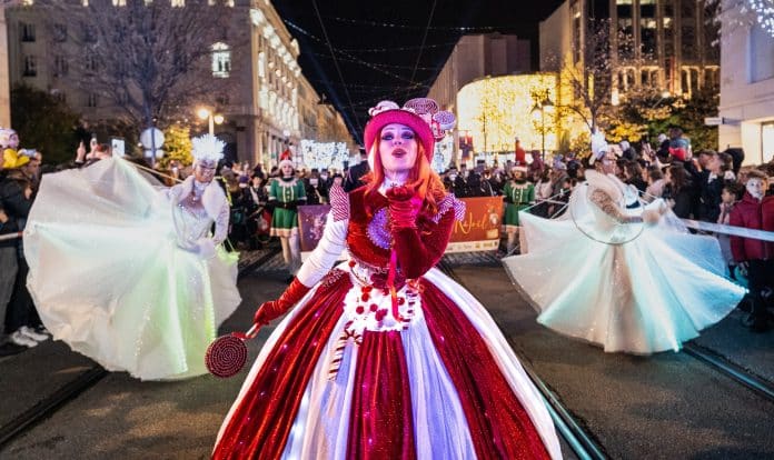 La Grande Parade de Noël de Nice Une dame déguisée en princesse avec de cheveux rouges fait un bisou devant la caméra avec des figurants derrière elle. La photo se déroule de nuit avec les décorations de Noël.