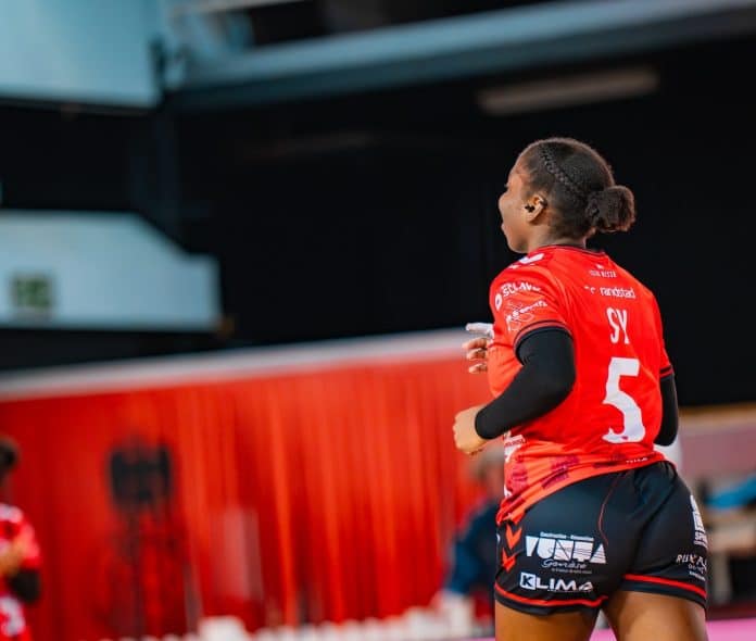 Une joueuse de l'OGC Nice handball en maillot rouge et short noir de dos sur le parquet.