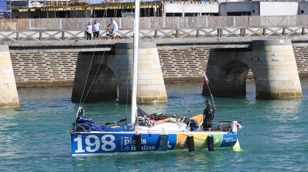 Nathan Mesiano sur son bateau bleu et blanc avec des touche de couleur orange, jaune et verte à l'arrière et le numéro 198 inscrit en gros à l'avant.