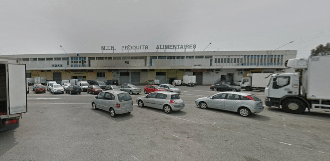 Le marché d'intérêt national de Nice avec un parking, des voitures, des camions devant un vieux bâtiment en taule.