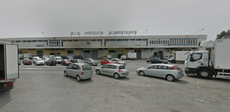Le marché d'intérêt national de Nice avec un parking, des voitures, des camions devant un vieux bâtiment en taule.