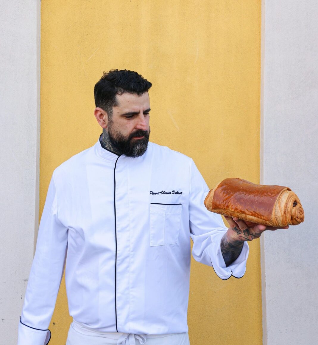 Le chef Pierre Olivier Dubost tient un pain au chocolat un grand pain au chocolat dans la main en le regardant sur un fond jaune.