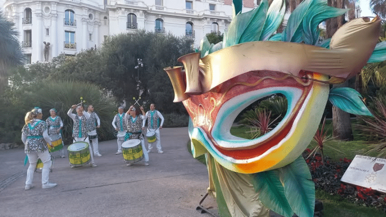 masque géant et coloré avec des feuilles du carnaval de nice. des personnes en tenues carnavalesque qui jouent du tambour à côté.
