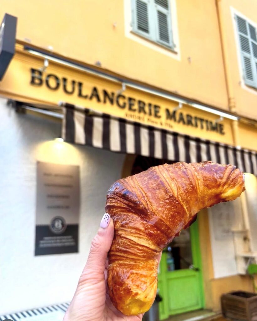 Un croissant tenu dans une main devant la devanture ancienne de la boulangerie Maritime à Nice.