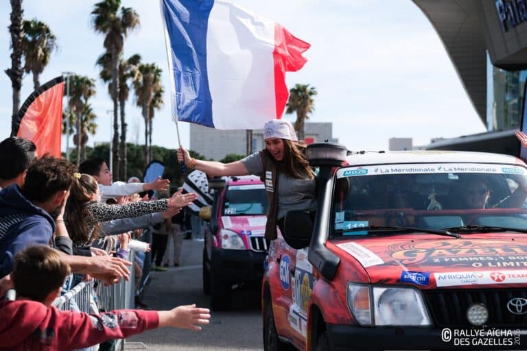 Une participante du rallye des gazelles sort par la fenêtre de sa voiture avec un drapeau français pour saluer la foule