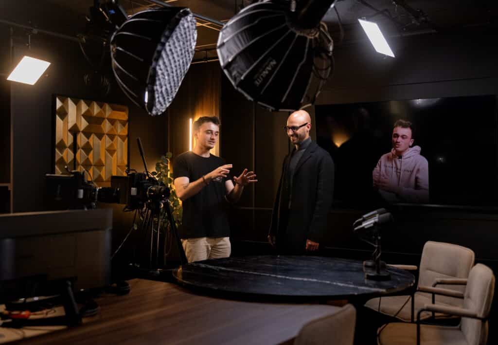 L'un des fondateurs présente le studio à des invités. On y trouve des spots lumineux, une grande table marron et du matériel de tournage.