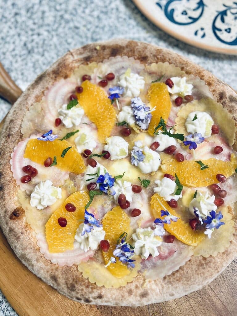 Une pizza aux couleurs de la méditerranée avec de la grenade, un fromage fondu et des tranches jaunes.