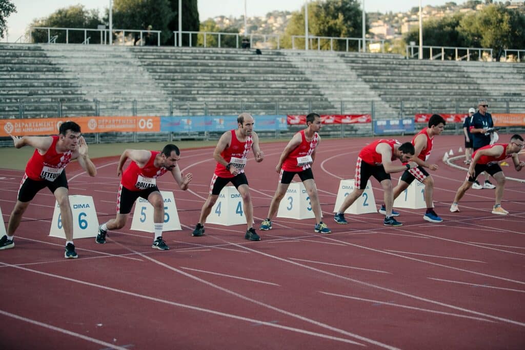 Des athlètes handisport, sur la ligne de départ devant leurs plots respectifs, en maillot rouge et short noir prêt à partir.