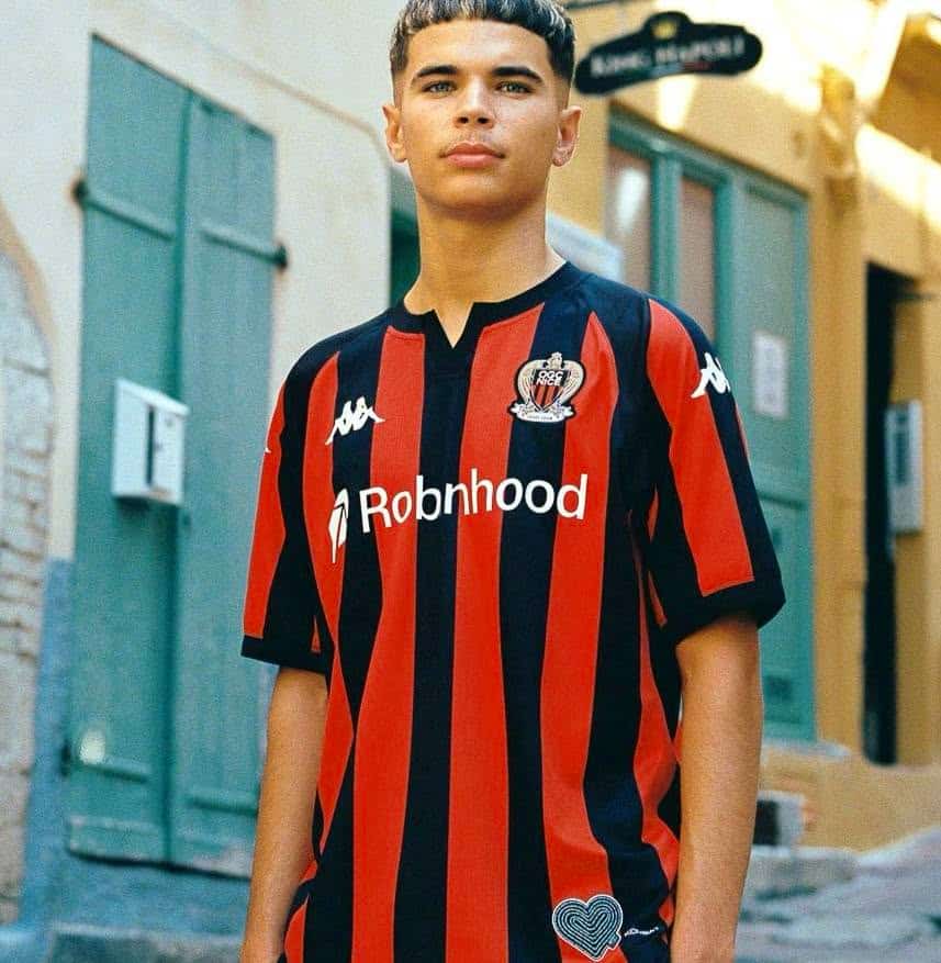Un jeune pour le maillot rouge et noir de l'OGC Nice pour la saison 2026-2027 très similaire aux années précédentes mais avec un couer en bas à gauche pour symboliser le soutient du club aux victimes de l'attentat du 14 juillet 2016 sur la Promenade des Anglais.