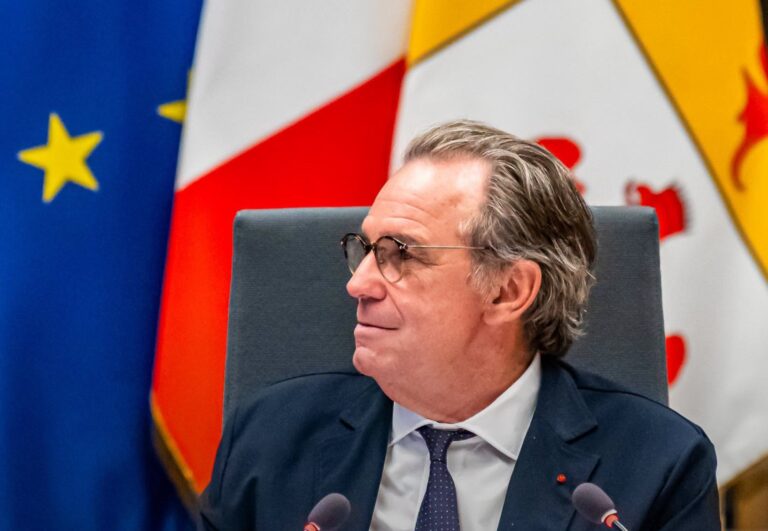 Le président de la Région SUD, Renaud Muselier regarde vers la droite avec des micros devant lui et les drapeaux de la région et de l'Europe derrière lui.