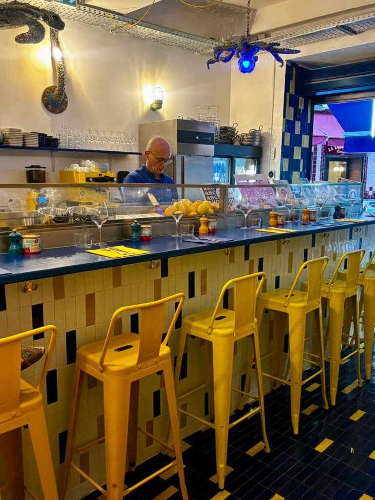 Le comptoir du restaurant Mer à Nice avec une ambiance ancienne, des tabourets jaunes, un comptoir en mosaïque jaune, bleu et rose pale.