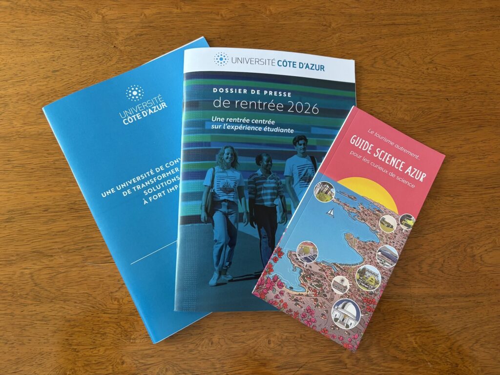 Trois brochures de l’Université Côte d’Azur posées sur une table en bois : une plaquette bleue institutionnelle, le dossier de presse de la rentrée 2026 avec trois étudiants en couverture, et le « Guide Science Azur » illustré.