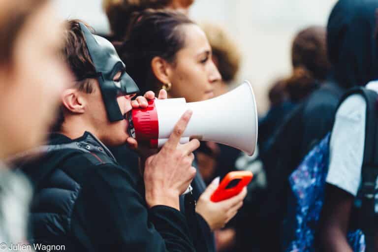 Un jeune avec un masque et un mégaphone parle à des personnes devant lui.